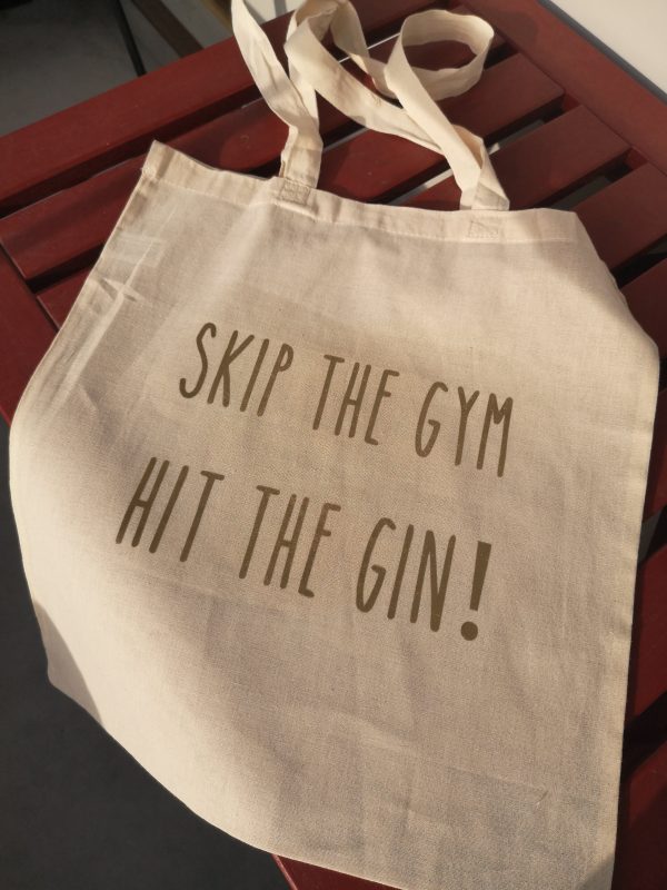 Natural tote bag with quote Skip the Gym, Hit the Gin! lying on red table.
